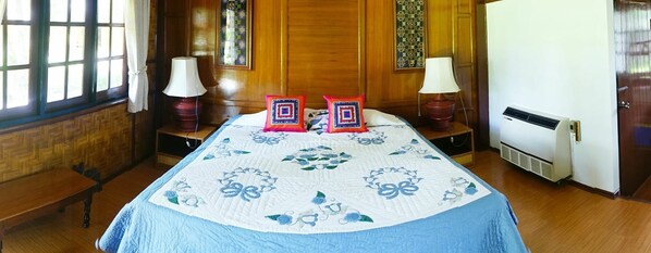 Habitación de hotel con piso de madera, cama con colcha estampada, dos lámparas de noche y una pared con vitral.