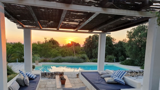 Ein überdachter Sitzbereich im Freien mit Pool, Steinmauern und Blick auf den Sonnenuntergang.