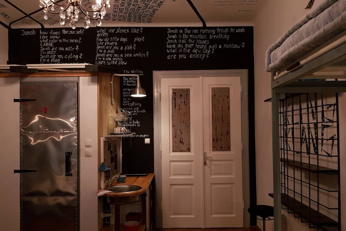 Una cocina compacta con un refrigerador de acero inoxidable, una encimera de madera y un lavamanos. Hay dos puertas blancas con acabado rústico y una escalera con barandillas de metal.