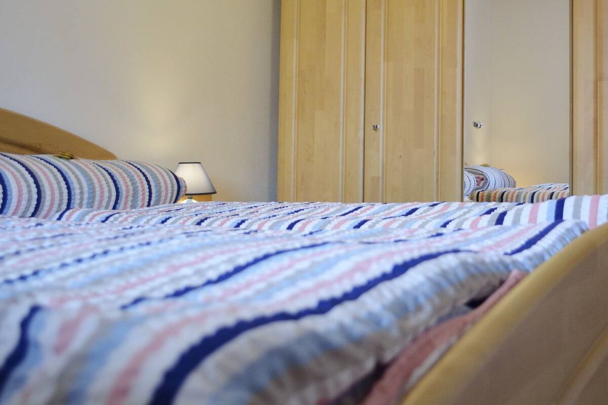 A bed with a striped bedspread, a bedside lamp, and a wooden wardrobe in the background.