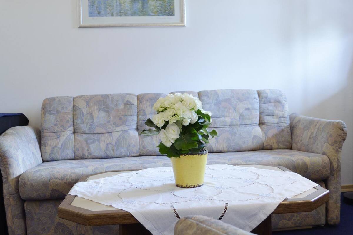 A living room with a patterned sofa, a small wooden table with a lace tablecloth, and a yellow vase with white flowers.