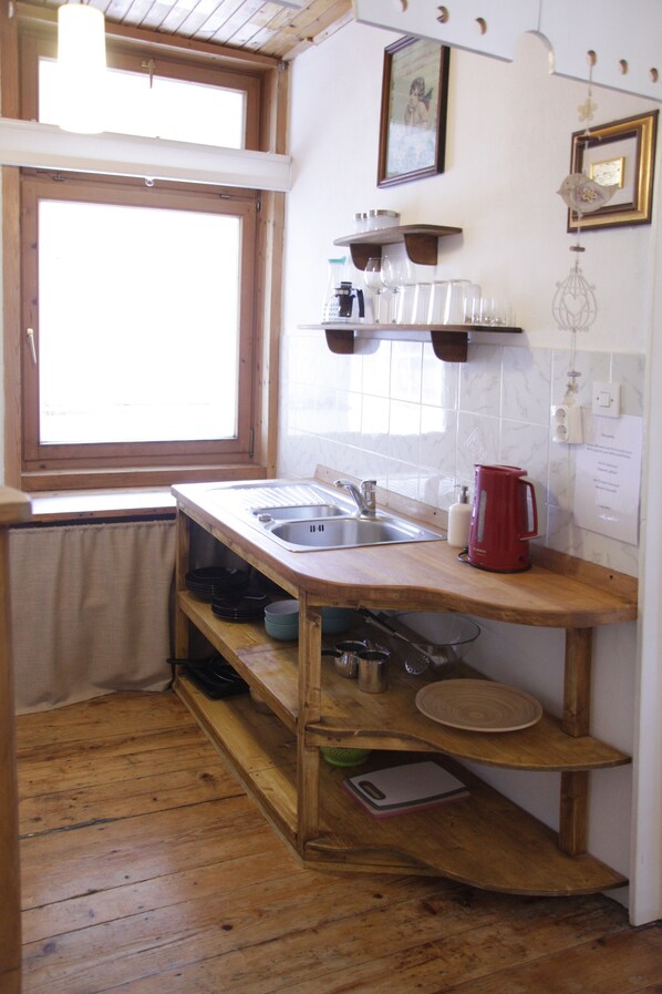 Una pequeña cocina con cubierta de madera, un lavamanos y una ventana. Hay repisas sobre la cubierta y una lámpara colgante.