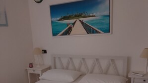 Habitación de hotel con cama, ropa de cama blanca, un cuadro de una playa y una mesita de noche con lámpara.