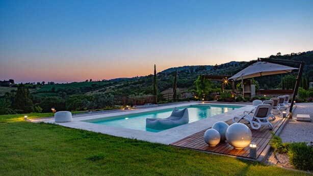 Una piscina con sillones de descanso y una pérgola, rodeada de césped y colinas al fondo.