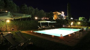 Area piscina notturna con poltrone, ombrelloni e un patio ben illuminato.