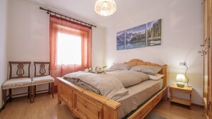 A bedroom with a wooden bed, a bench, a window with curtains, a wall-mounted lamp, and two framed pictures. - Martina Ski in - Ski out 50m, Livigno, Italy (Livigno)