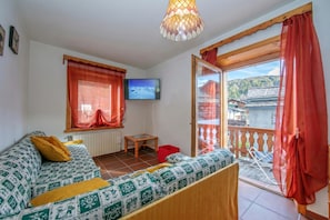 A room with a sofa, a television, a wooden table, and a balcony with a view of a building and trees. - Martina Ski in - Ski out 50m, Livigno, Italy (Livigno)