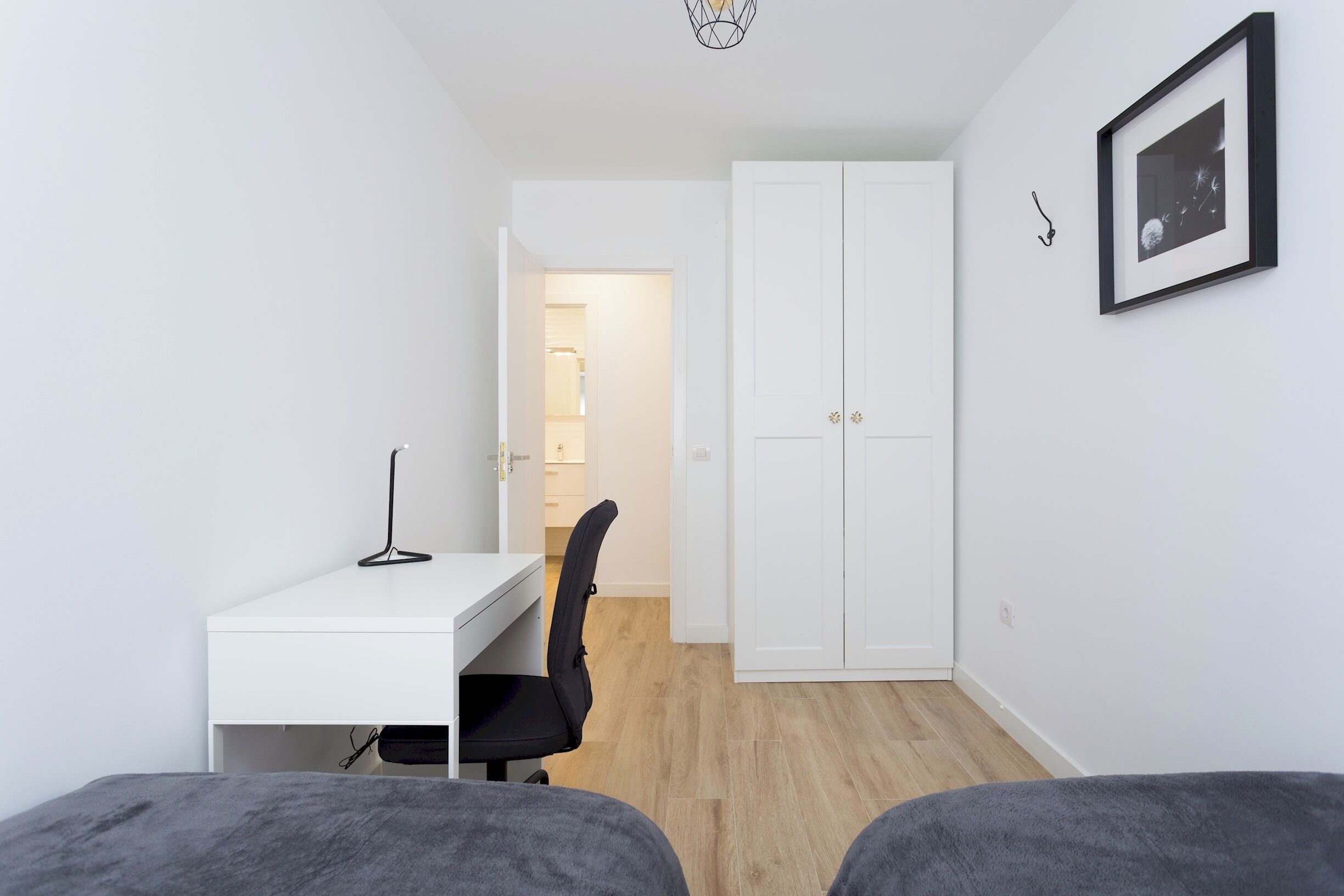 A small, well-lit room with a white desk, a black office chair, a wardrobe, and a framed picture on the wall.