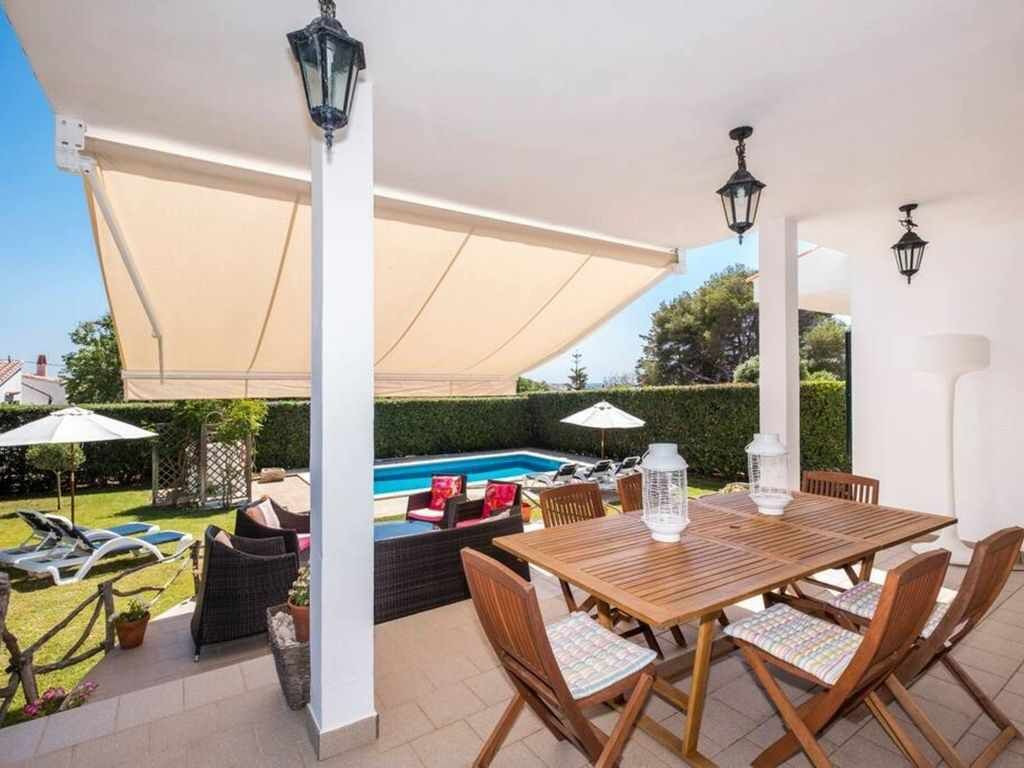 A covered patio area with a pool, wooden table, and chairs, surrounded by greenery and a white fence.