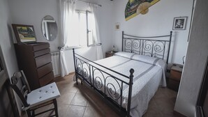 Un dormitori amb un llit, una petita taula de nit, una cadira, una finestra amb cortines i una paret amb ventilador i quadres.