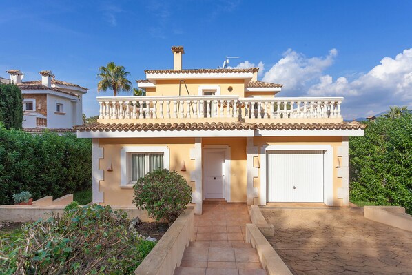 Une maison à deux étages, avec un toit recouvert de tuiles, un balcon doté de balustrades blanches et une porte d’entrée ornée d’une petite fenêtre.