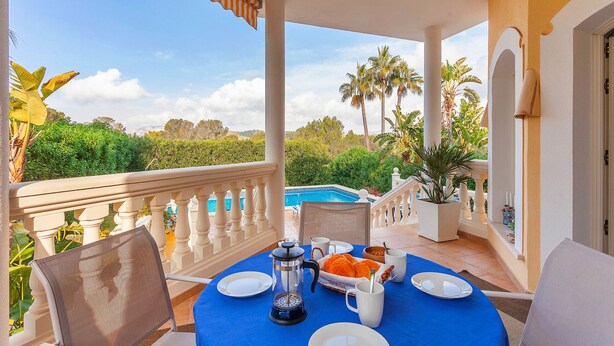 A balcony with a table set for breakfast, overlooking a pool and lush greenery.