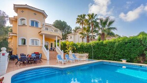 A pool area with lounge chairs and a dining set, surrounded by a well-maintained garden and a two-story building with balconies.