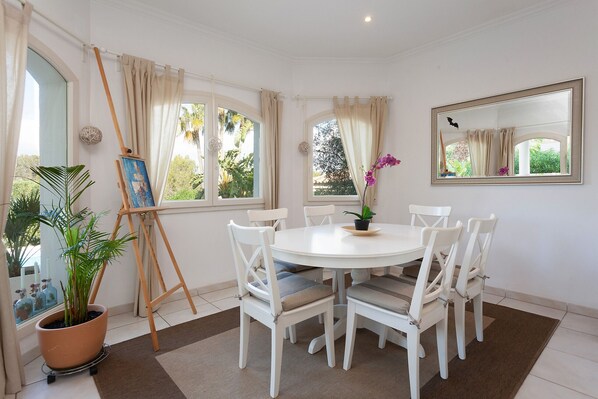 A dining area with a round table, white chairs, a potted plant, and a painting on an easel.