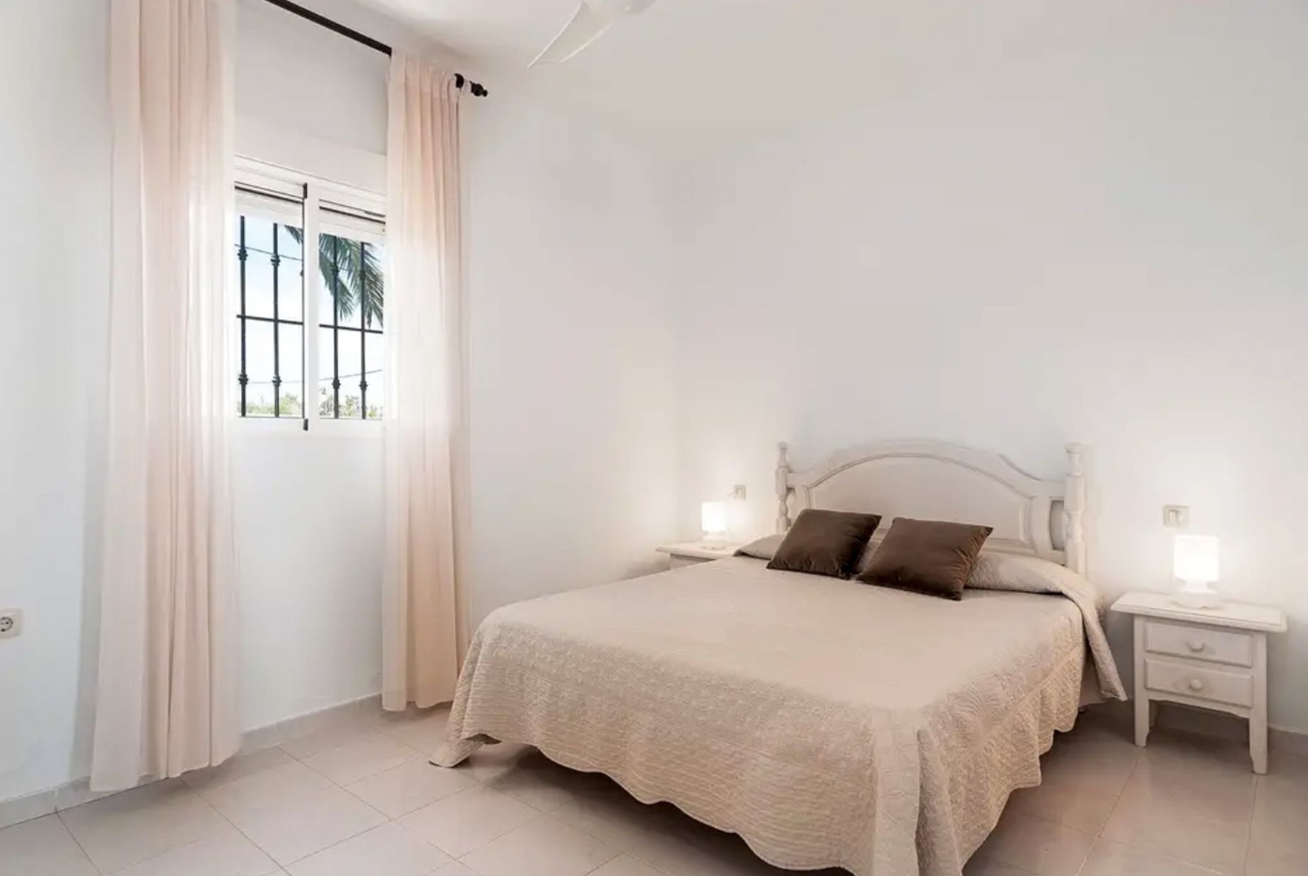 A bedroom with a bed, two bedside tables, a window with curtains, and a view of palm trees.