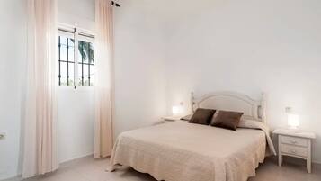 A bedroom with a bed, two bedside tables, a window with curtains, and a view of palm trees.