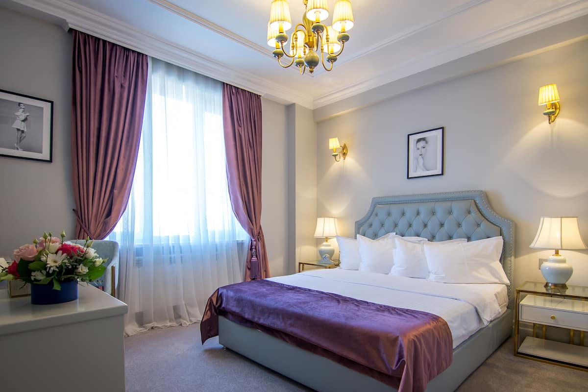 Ein Hotelzimmer mit einem großen Bett, zwei Nachttischen mit Lampen, einer Kristallleuchtre und einem Fenster mit Vorhängen.