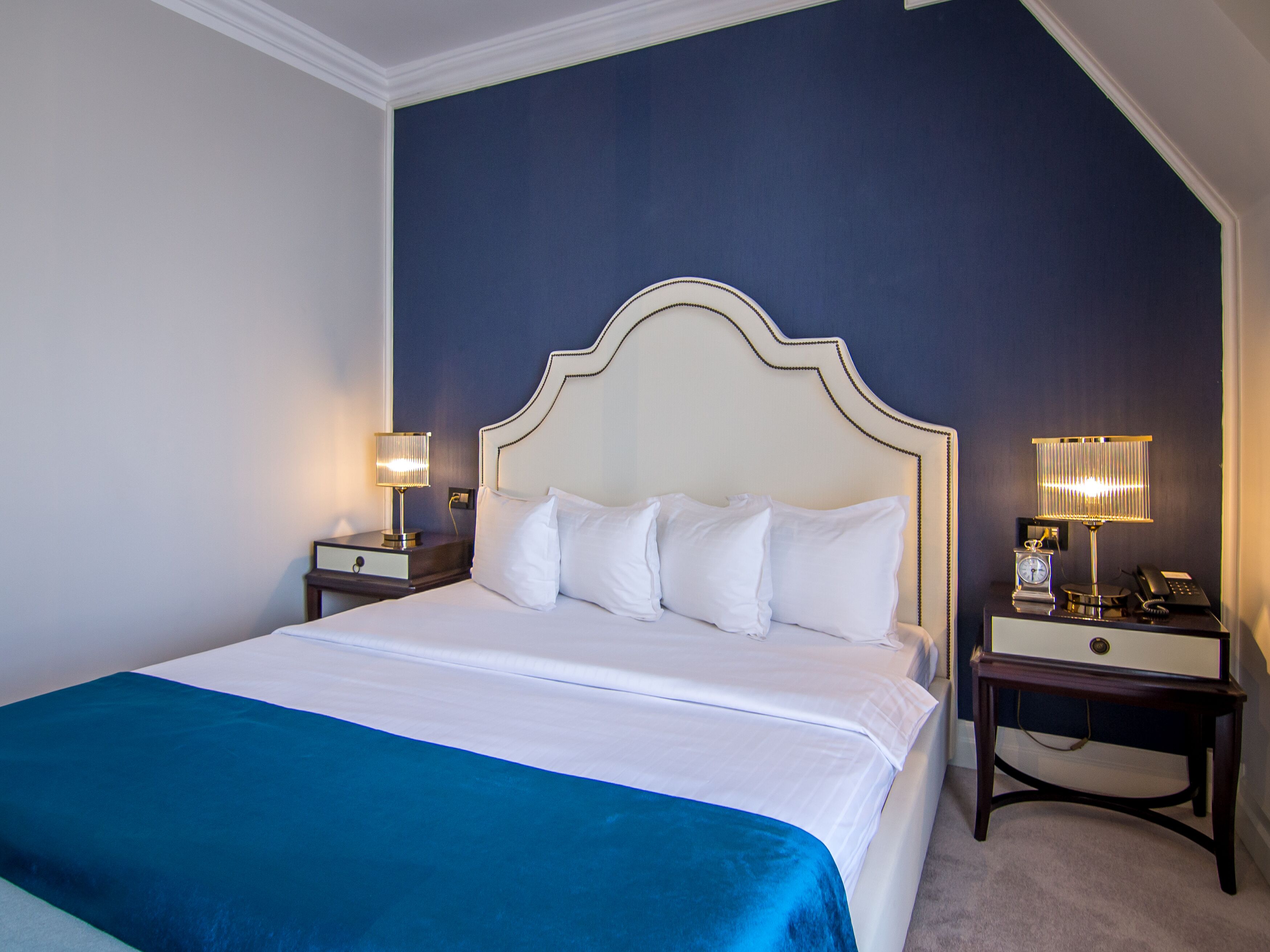 Una cama bien hecha con ropa de cama blanca y un detalle azul, dos mesitas de noche con lámparas, y una pared azul oscuro.