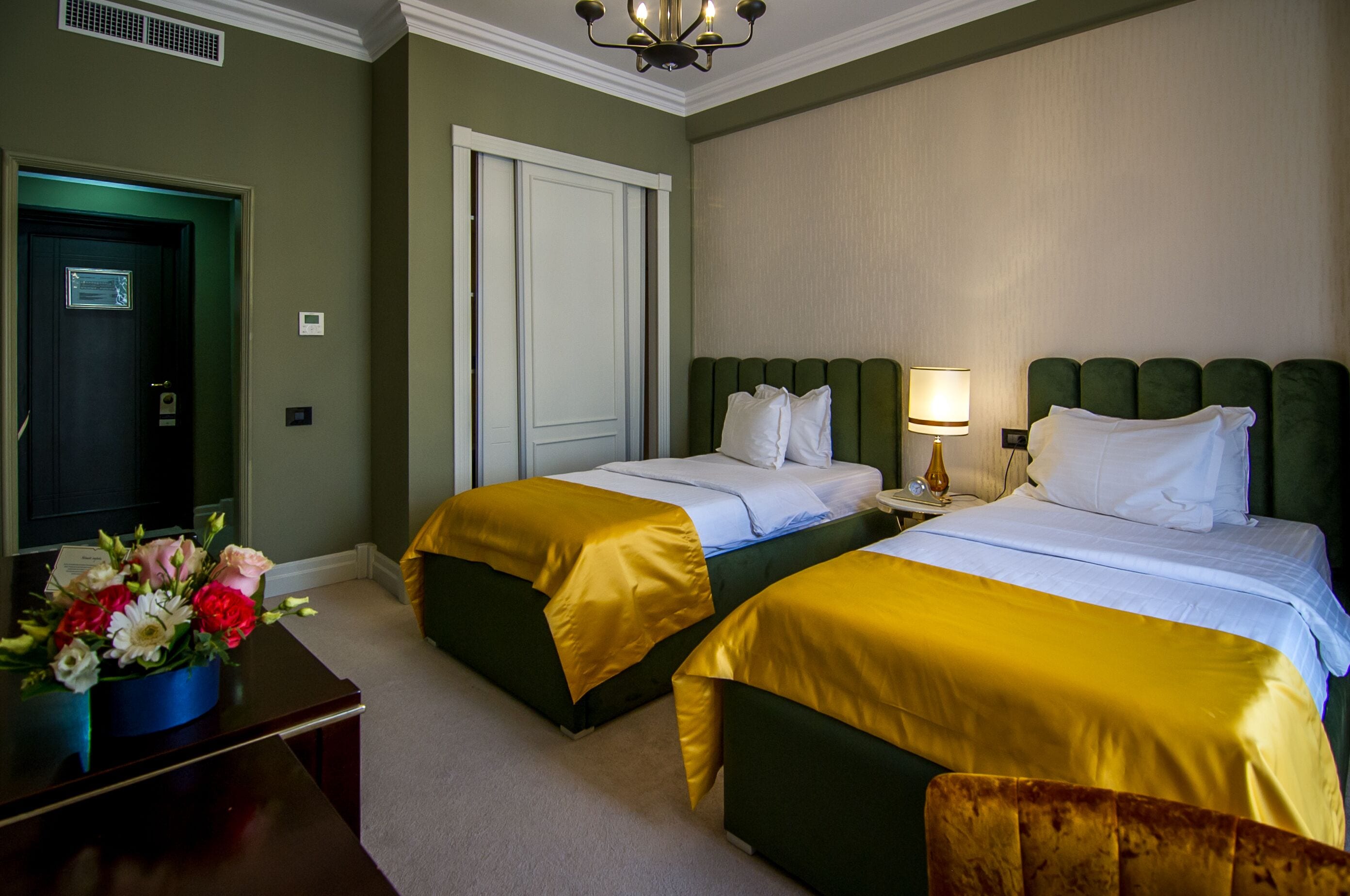 Ein Hotelzimmer mit zwei Betten, einem Schreibtisch mit Blumenvase und einer Tür mit Lampe darüber.