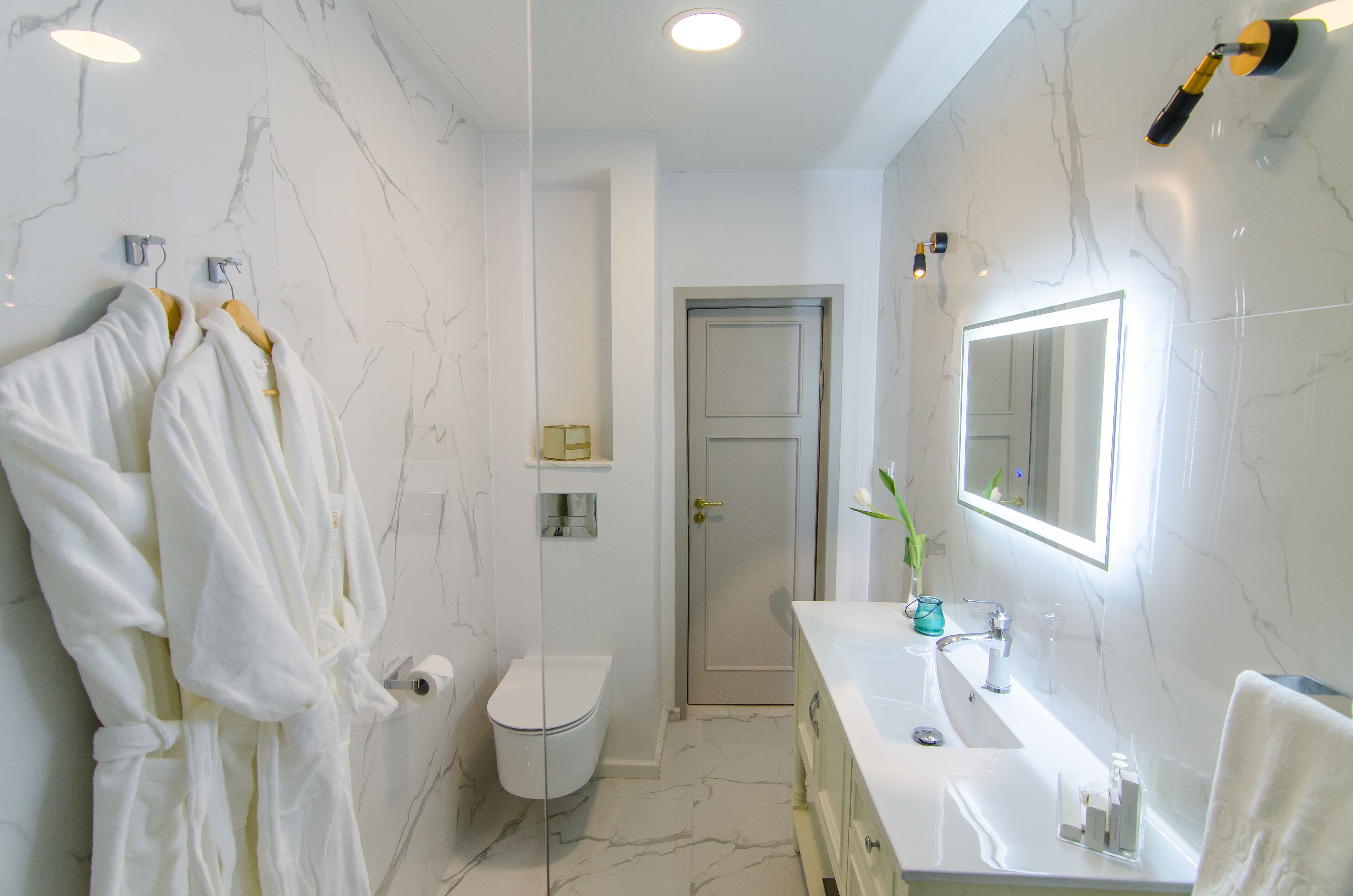 Un baño con pared de mármol, un lavabo blanco, un espejo y dos albornoces blancos colgados en un perchero.