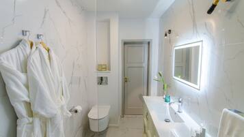 Un baño con pared de mármol, un lavabo blanco, un espejo y dos albornoces blancos colgados en un perchero.