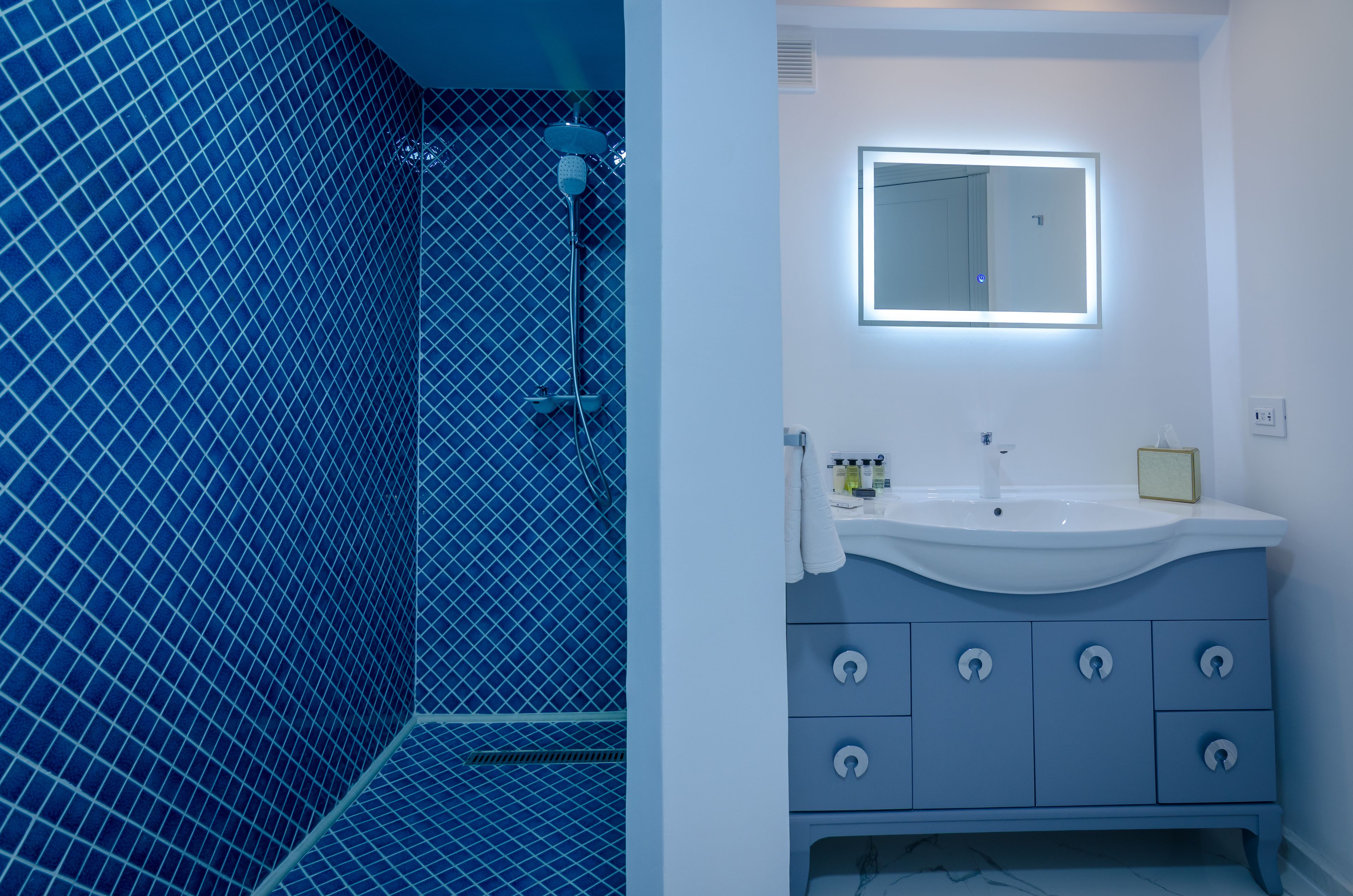 A bathroom with a blue tiled shower, a white sink with a mirror, and a vanity with drawers.