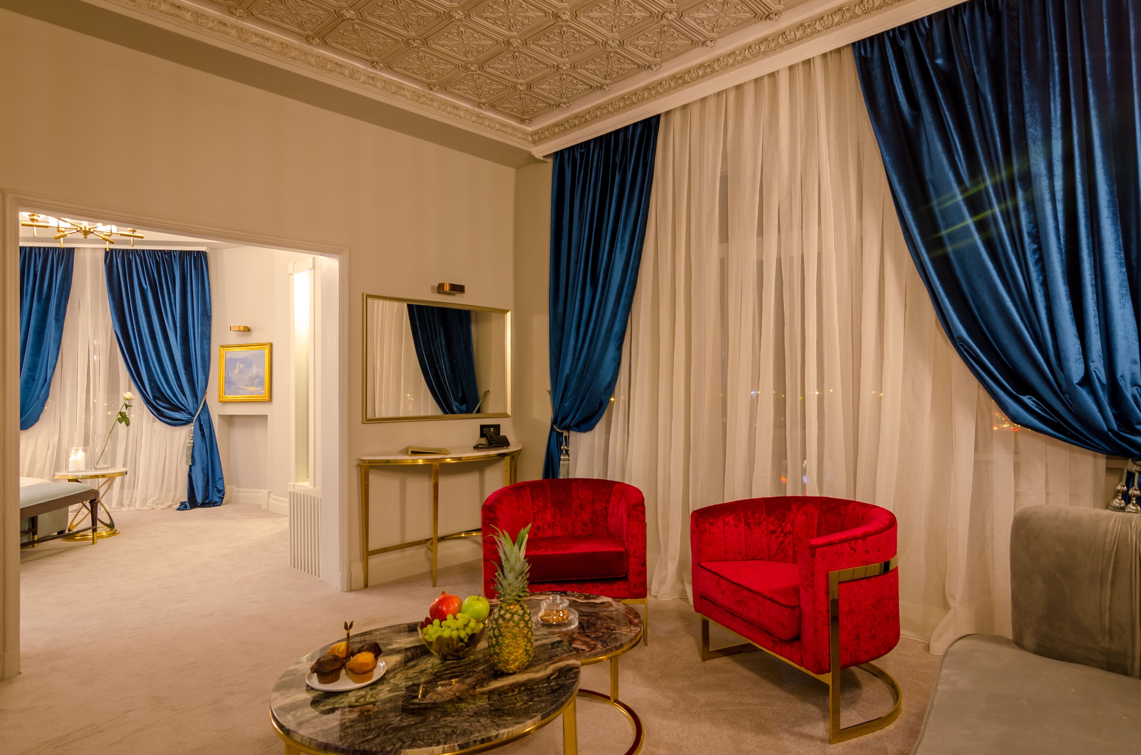A room with a marble coffee table, red velvet armchairs, a mirror, and blue curtains.