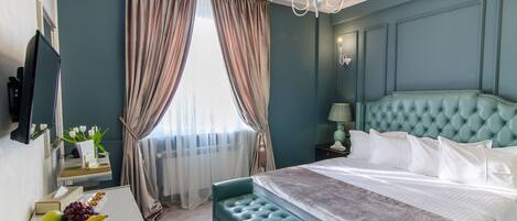A bedroom with a tufted headboard, a large bed, a desk with a chair, a television, and a window with curtains.