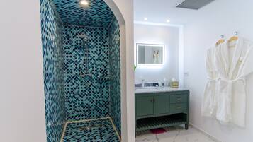 Un baño con ducha de azulejos azules, un tocador verde con espejo y dos batas blancas colgadas en la pared.