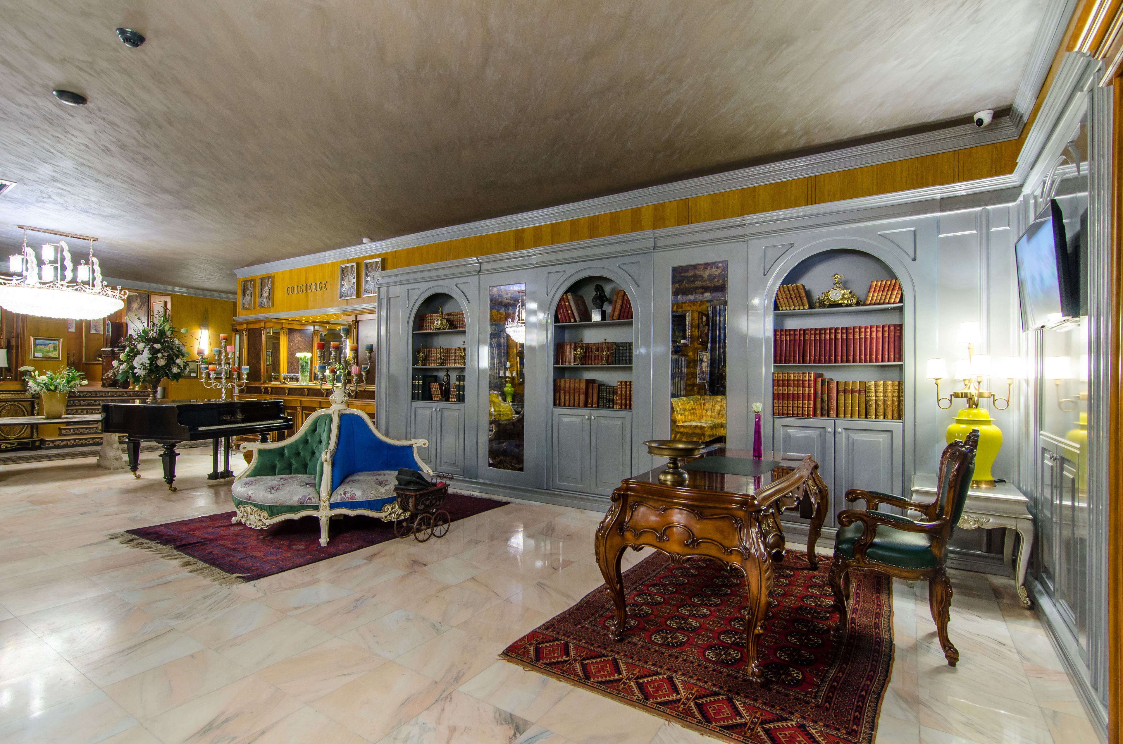 A grand lobby with a piano, a blue armchair, a wooden table, and a bookshelf filled with books.