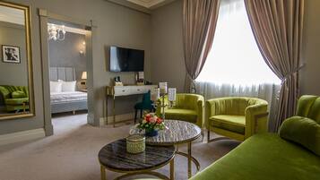 A hotel room with a bed, a desk with a flat-screen TV, two green armchairs, a round coffee table, and a large window with curtains.