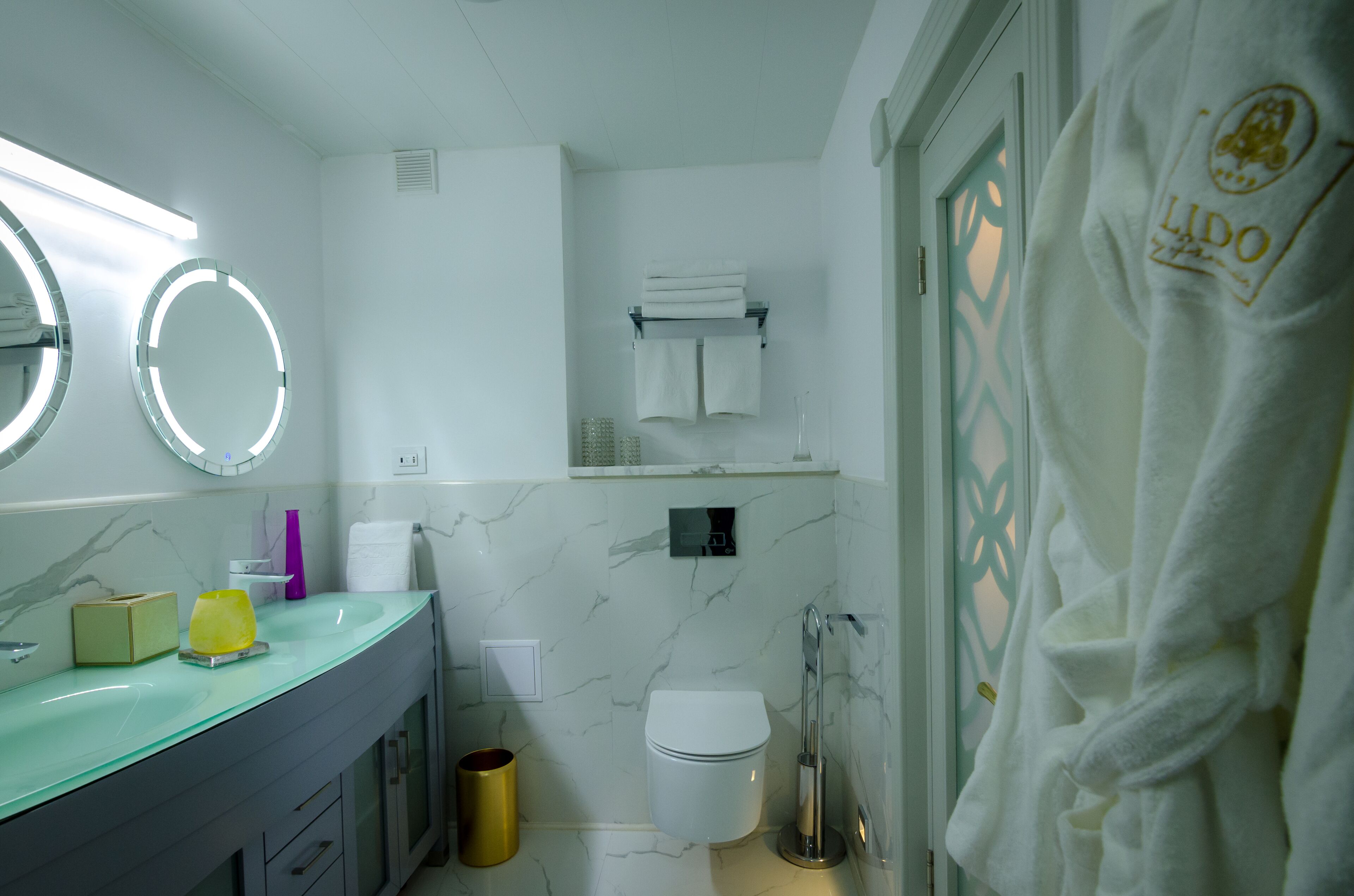 A modern bathroom with a marble wall, a glass sink, a toilet, and a towel rack.