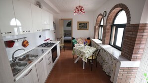 Une cuisine compacte avec des meubles blancs, une cuisinière à gaz et un évier. On y trouve un coin repas avec une table et des chaises, ainsi qu’une fenêtre ornée d’un motif en briques.