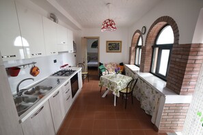 Une cuisine compacte avec des meubles blancs, une cuisinière à gaz et un évier. On y trouve un coin repas avec une table et des chaises, ainsi qu’une fenêtre ornée d’un motif en briques.