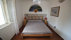 Une chambre avec un lit en bois, une fenêtre avec des rideaux blancs, un ventilateur fixé au mur, un tableau et une photographie encadrée.