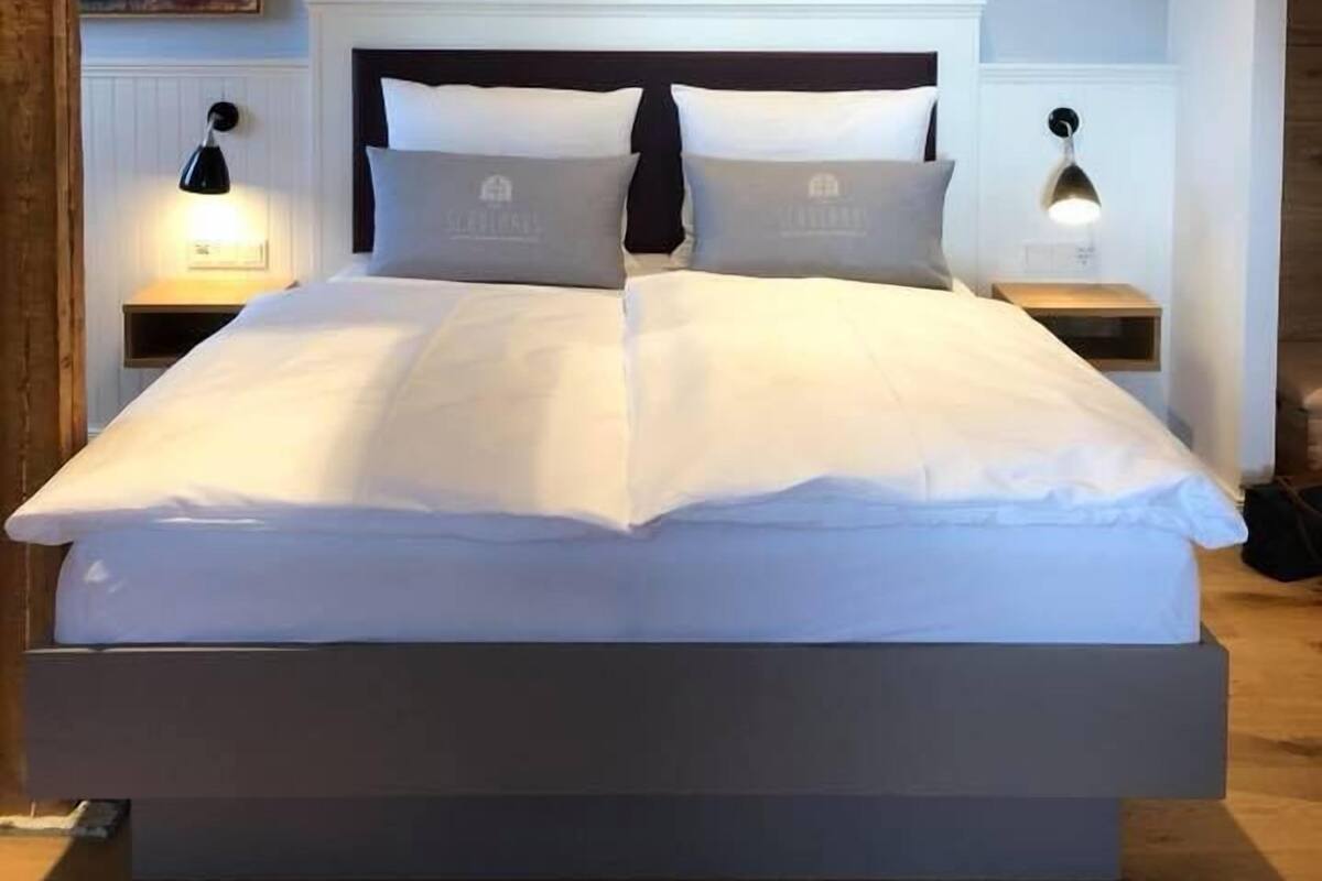 Quarto moderno com uma cama grande, vigas de madeira, paredes brancas e duas luminárias de cabeceira.