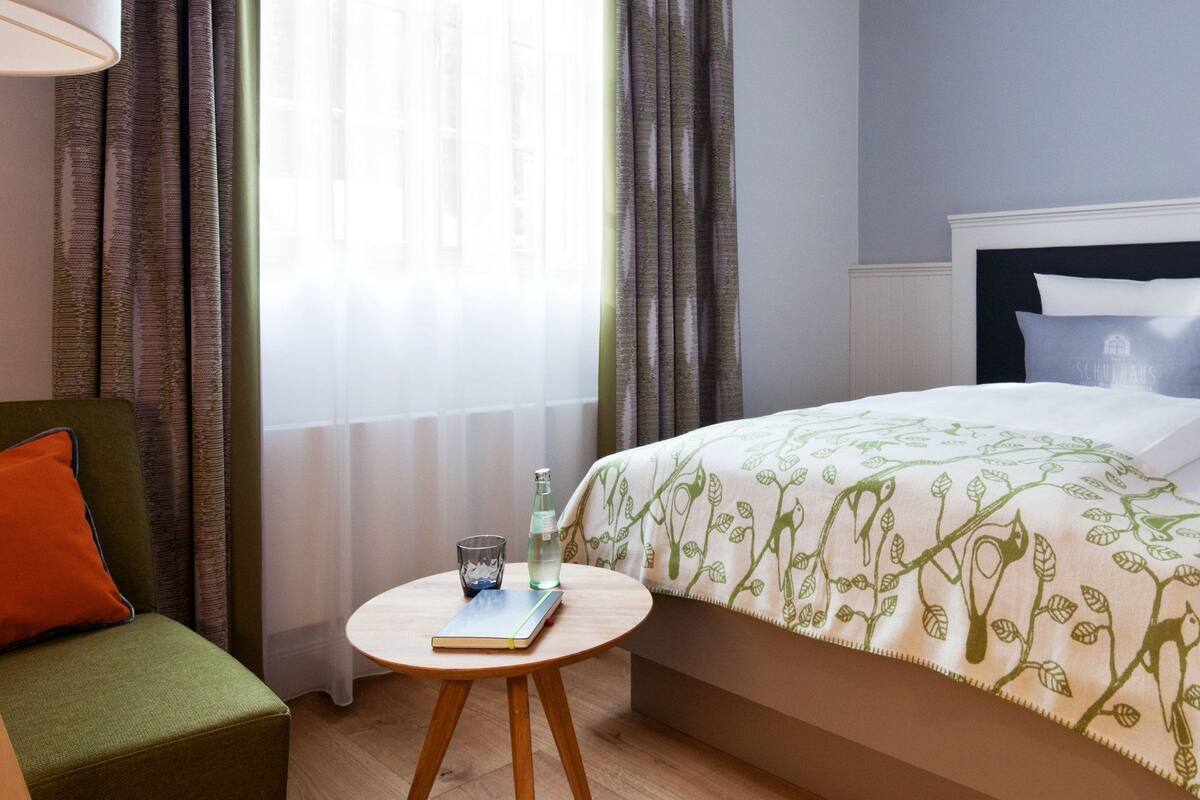 Quarto de hotel com cama, uma cadeira, uma pequena mesa redonda com um livro e uma garrafa, e uma janela com cortinas.