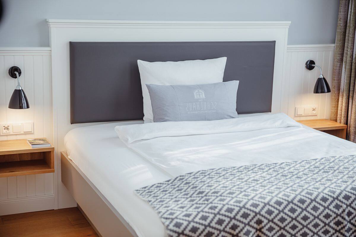 Cama bem arrumada com manta estampada, cabeceira branca e duas luminárias de apoio.