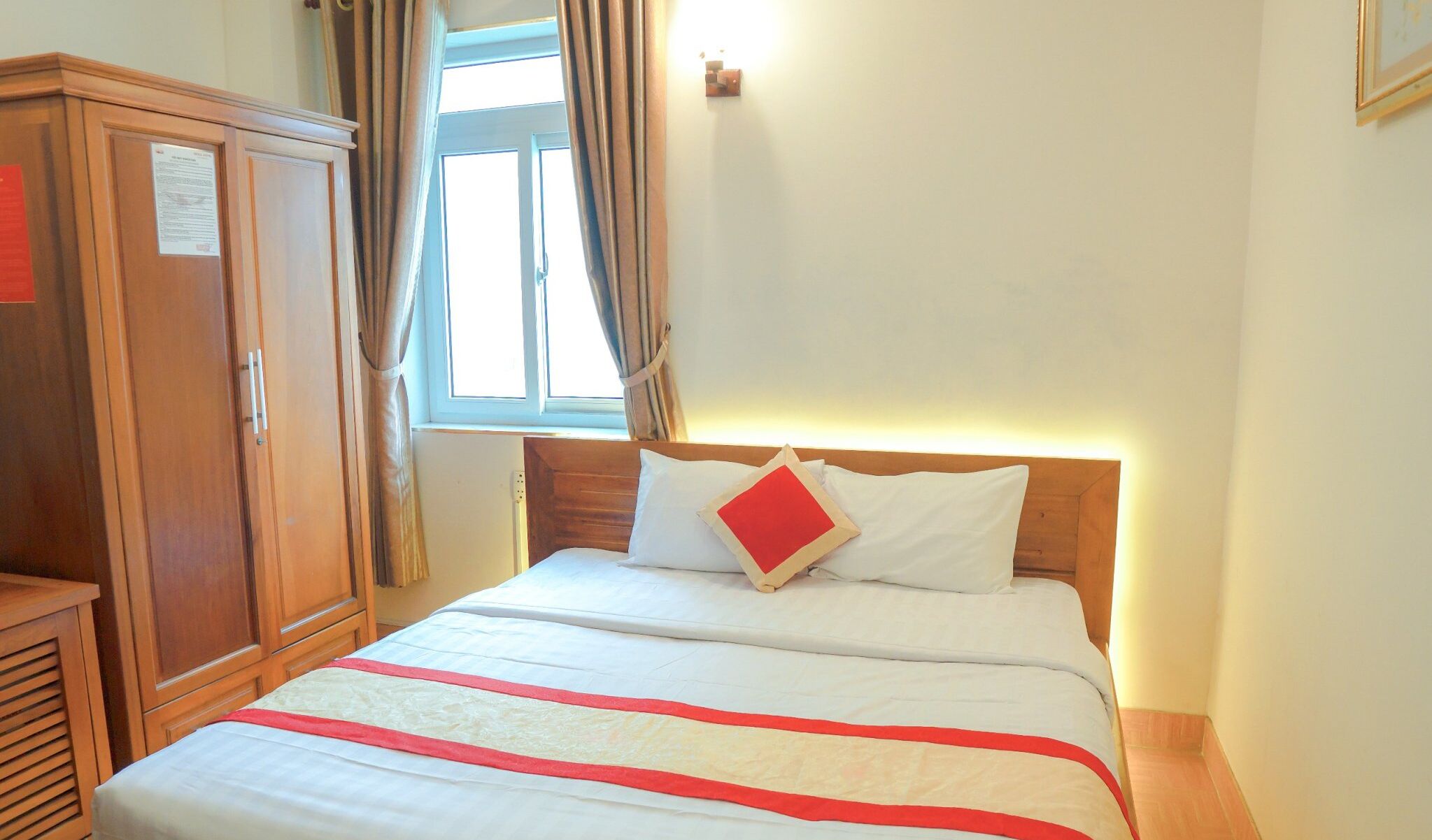 Une chambre d’hôtel avec une armoire en bois, un lit avec une literie blanche et un coussin rouge, et une fenêtre avec des rideaux.