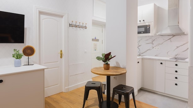 A modern kitchen with white cabinetry, a marble backsplash, and a built-in oven. There is a round wooden table with two stools, a small potted plant, and a wall-mounted light fixture. - Casa Armonía, charming apartment in the centre (Málaga)