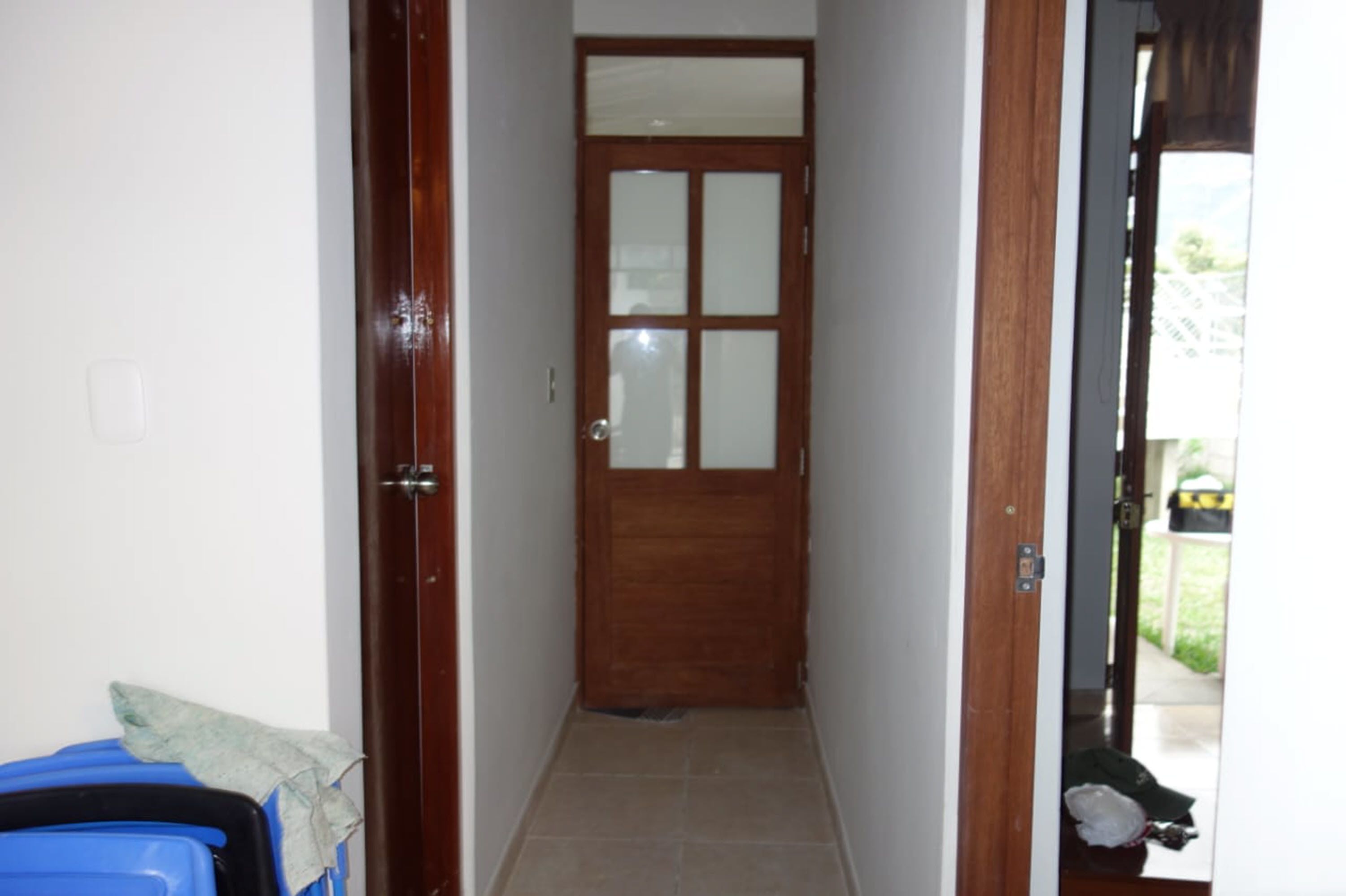 Un corridoio con una porta in legno, pavimento piastrellato e un cestino visibile.