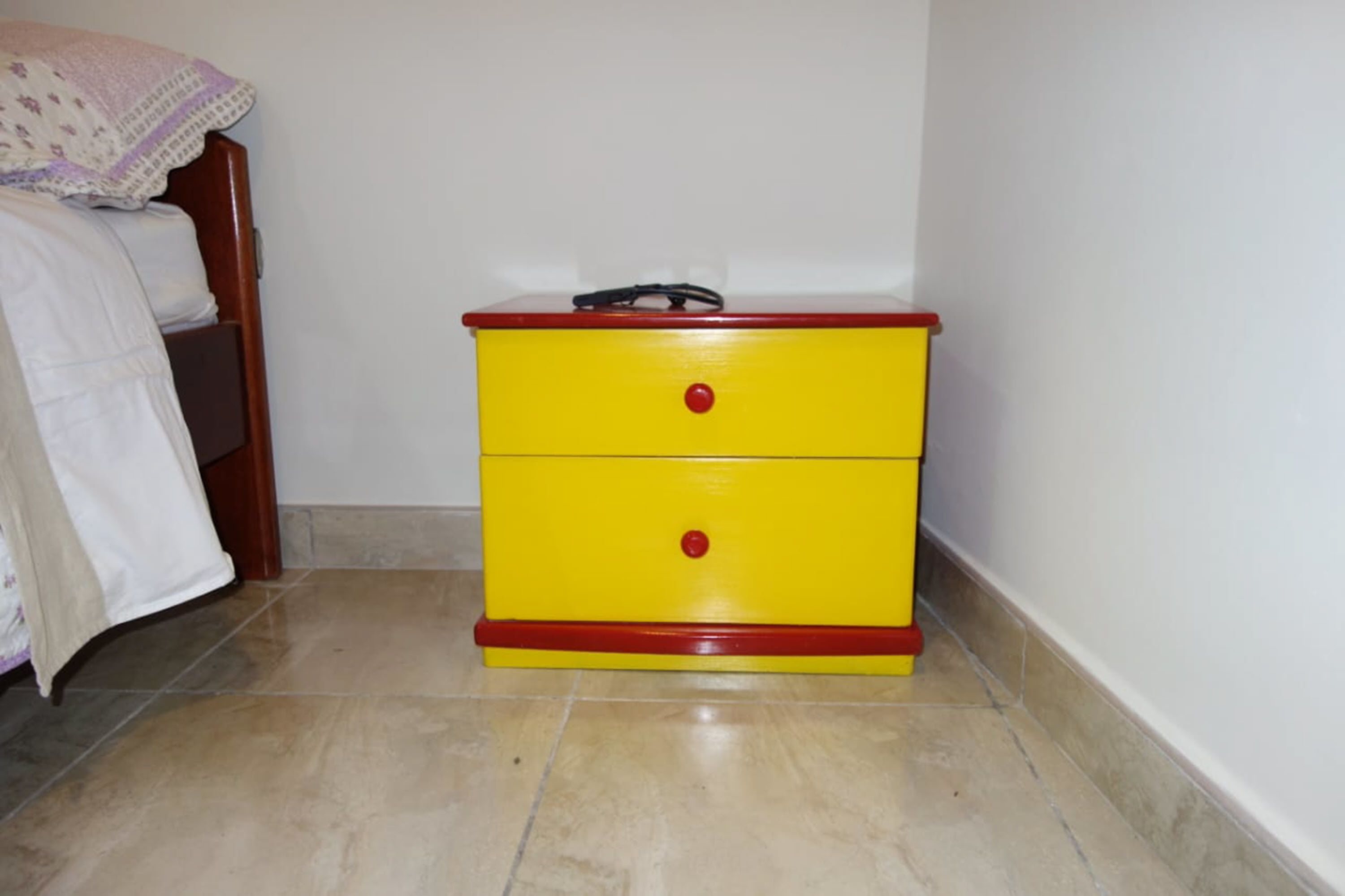 A yellow bedside table with two drawers and a red top, positioned next to a bed with a white blanket and a wooden headboard.