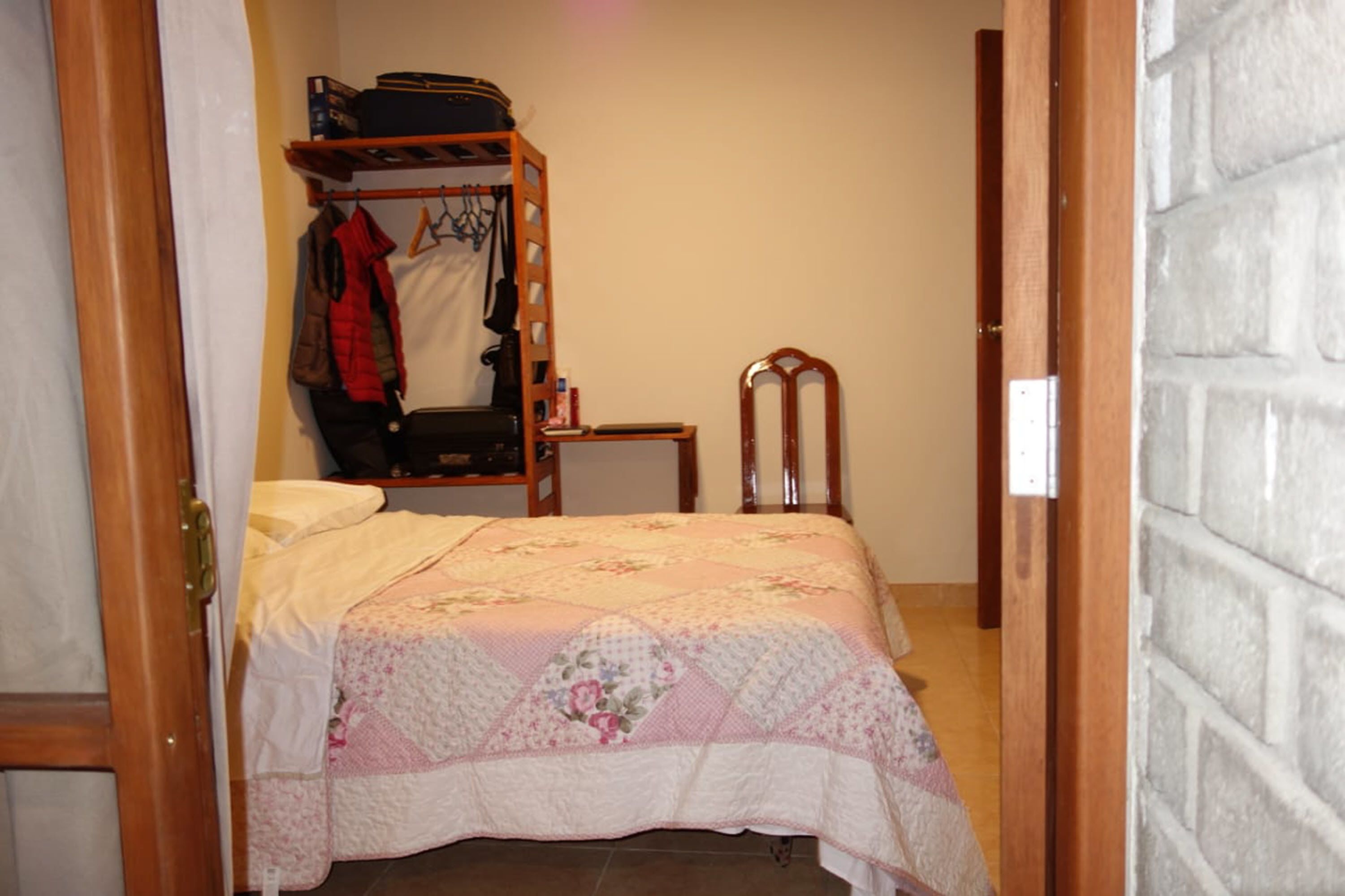 Un dormitori amb un llit, una cadira de fusta, un penjador amb roba i una maleta a la paret.