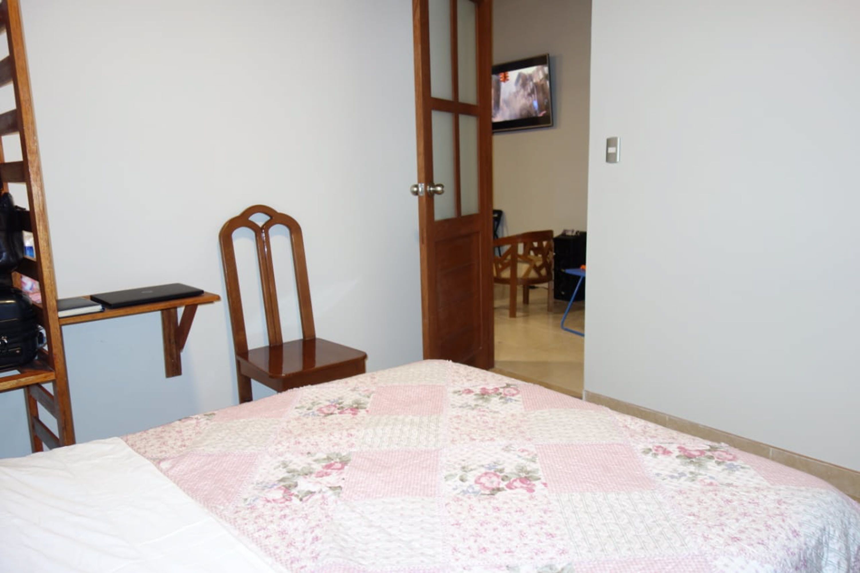 Un dormitorio con una cama, una silla de madera, un escritorio con una computadora portátil y un televisor en la pared.
