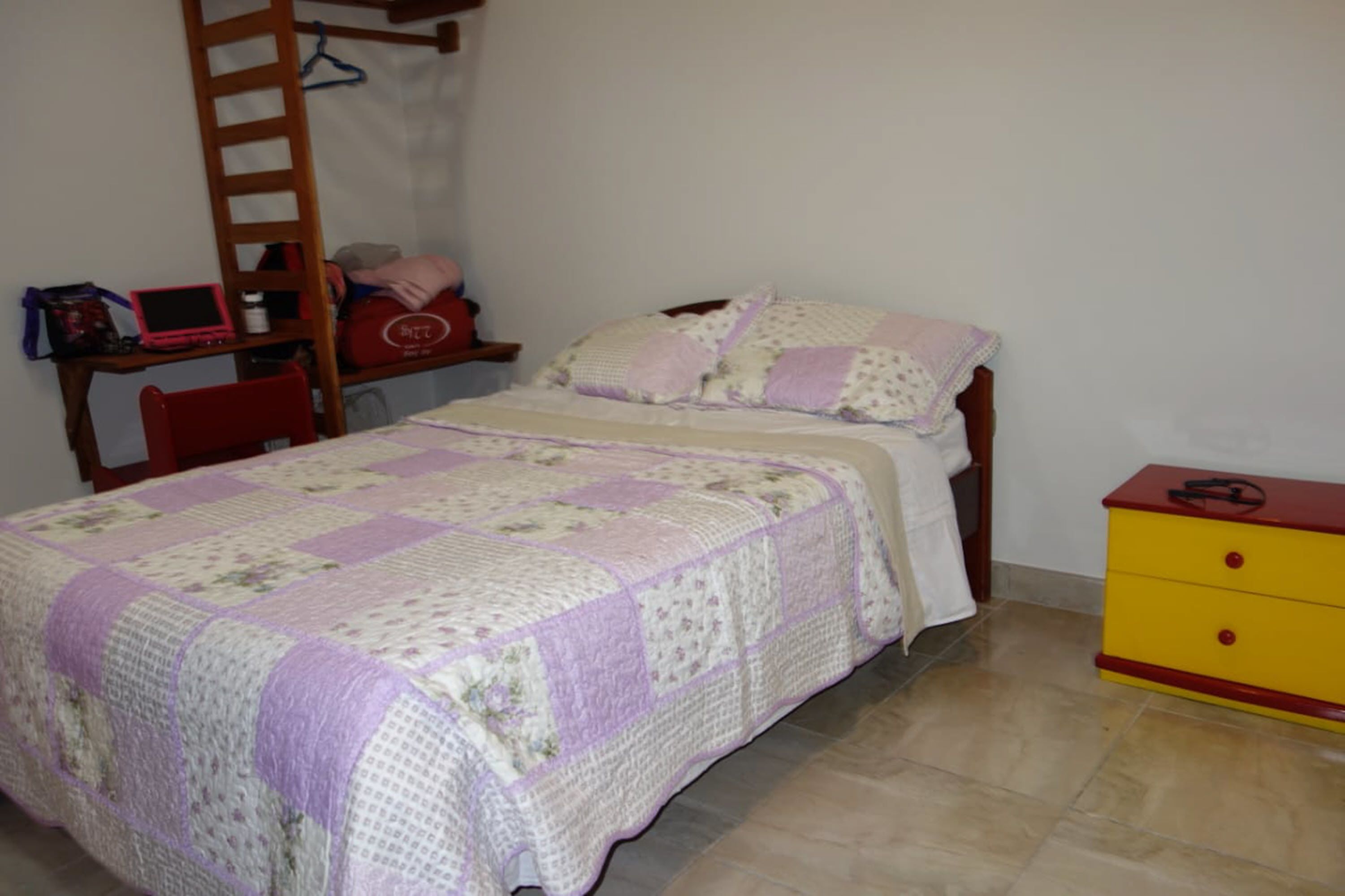 Un dormitorio con una cama, un escritorio con una computadora portátil, una silla, un aparador y una escalera apoyada en la pared.