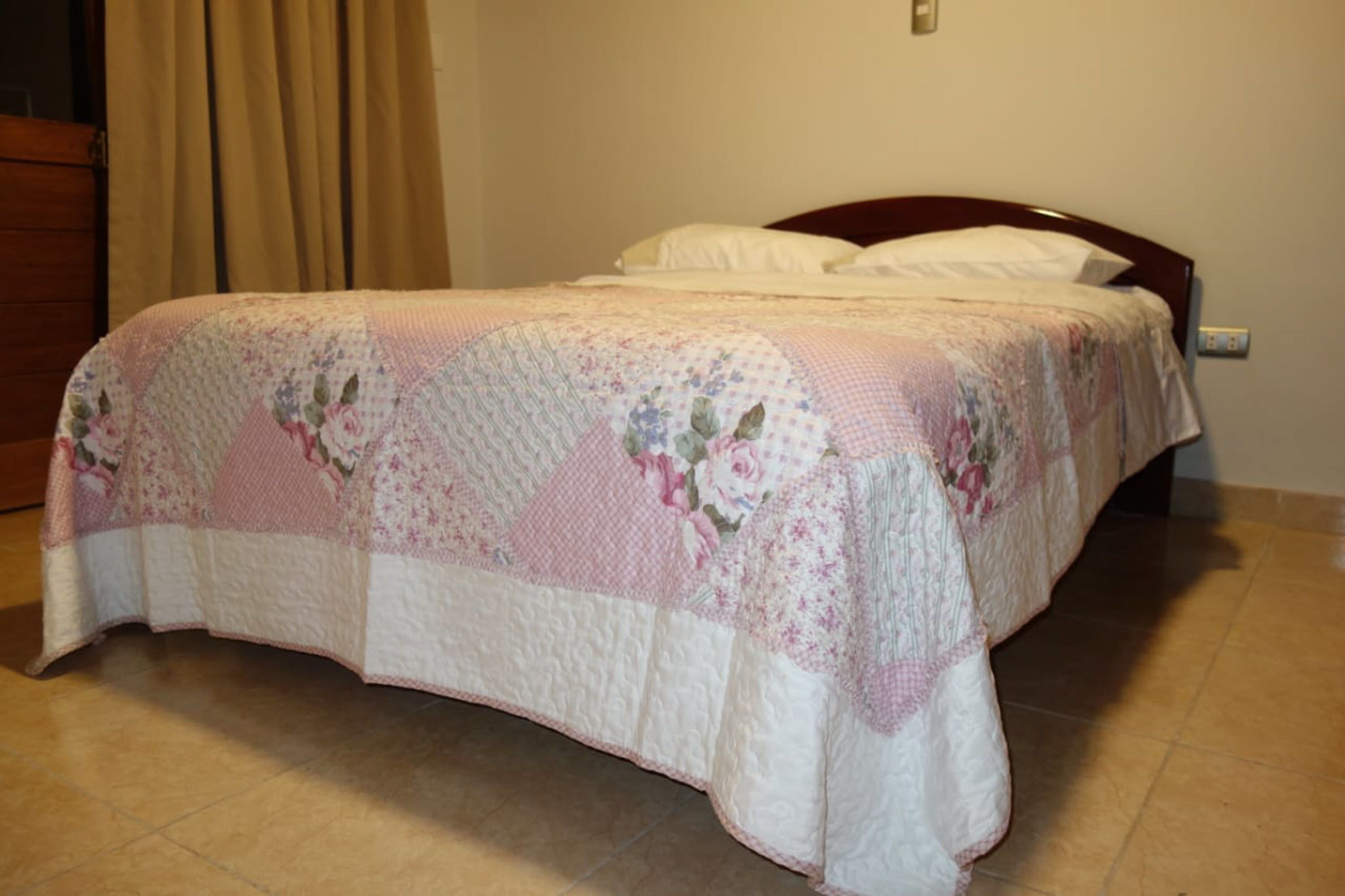 Un lit bien fait, recouvert d’une courtepointe à motifs floraux, avec une tête de lit en bois et un mur sobre agrémenté d’un interrupteur.