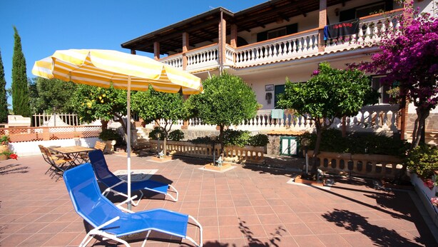 Ein Innenhof mit blauen Liegestühlen und einem gelb-gestreiften Sonnenschirm, umgeben von einer gefliesten Terrasse und einem Gebäude mit Balkon.