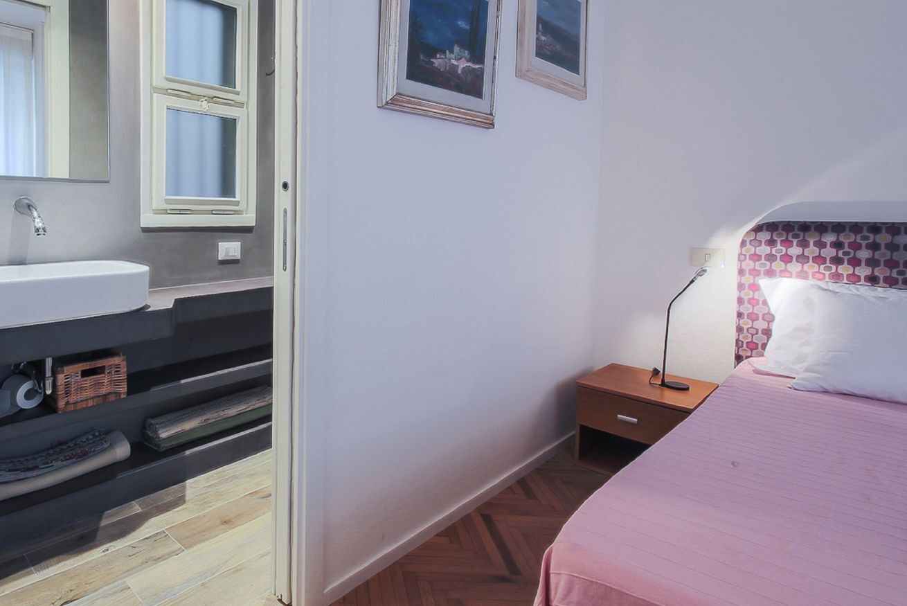 A bedroom with a bed, a nightstand, a lamp, a window with curtains, and a bathroom with a sink and mirror.