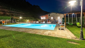 A swimming pool with lounge chairs and a swing set, surrounded by a stone building and a grassy area.
