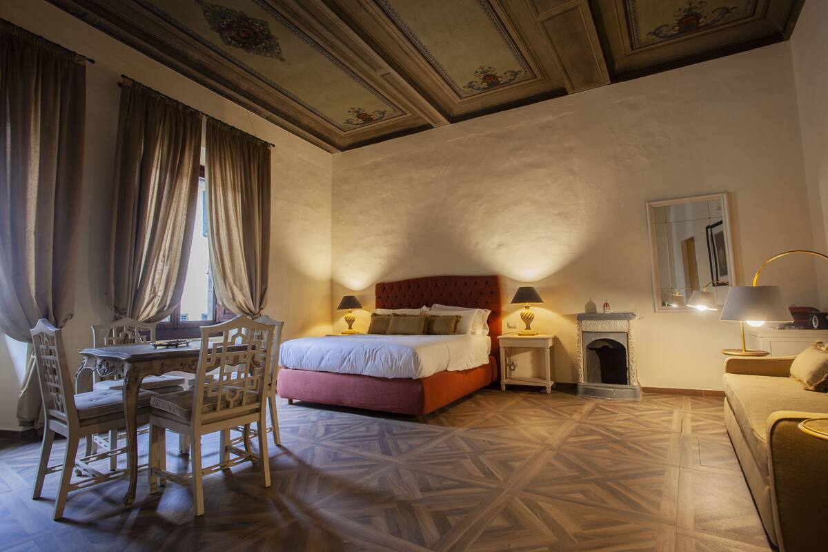 Een slaapkamer met een bed, nachtkastjes, een open haard, een eettafel met stoelen en een spiegel aan de muur.